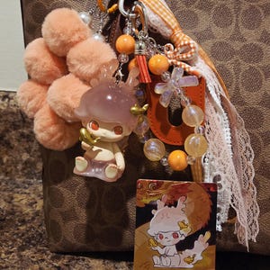 May include: A collection of decorative bag charms and accessories. The charms include a plush peach-colored pom-pom cluster, a small figurine, and a card with a cartoon illustration. Additional elements include orange beads, a tassel, and ribbons.