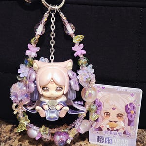 May include: A pink and purple charm featuring a cat-eared figurine surrounded by beaded accents and a small card. The charm has a silver chain and a variety of beads in shades of pink, purple, and green.