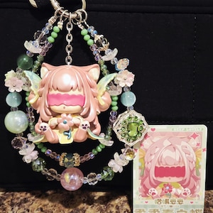 May include: A pink cat-eared figurine with a pink blindfold, adorned with beaded strands of green, clear, and pink beads, and a green gem pendant. A matching card with Japanese text is also present.