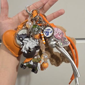 May include: A collection of keychains and charms in various shapes and colors. Includes a brown and white pony figurine, a heart-shaped orange leather charm, and several round charms with different designs. The keychains are attached to a silver metal clip and orange cord.