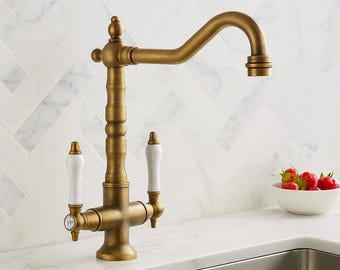 May include: A vintage-style kitchen faucet with a brass finish. It features white porcelain handles and a curved spout. The faucet is mounted on a white countertop with a marble tile backsplash. A bowl of strawberries sits nearby.