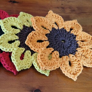 May include: A collection of handmade crochet flower coasters in various colors, including blue, red, yellow, orange, and black. These decorative coasters are arranged on a wooden surface, showcasing their vibrant hues and intricate petal designs.