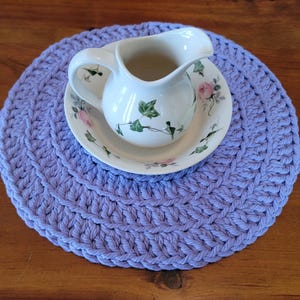 May include: A white ceramic pitcher and saucer set with floral and ivy designs sits on a round, lavender crocheted placemat. The pitcher has a curved handle and spout. The placemat is approximately 30 cm in diameter.