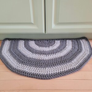 May include: A half-circle crochet rug in shades of gray and white. The rug features concentric rows of crochet stitches, creating a textured surface. The rug is placed on a wooden floor, with a light green cabinet in the background.
