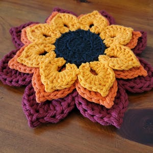 Può includere: Un sottobicchiere a forma di girasole fatto a mano all'uncinetto, con un centro blu scuro, circondato da strati di petali gialli, arancioni e bordeaux. Il sottobicchiere è realizzato con filato spesso e poggia su una superficie di legno. Il design a girasole.