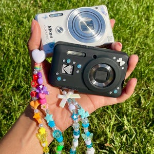 Puede incluir: Una cámara digital Nikon Coolpix blanca y una cámara digital Kodak negra, decoradas con pequeñas gemas adhesivas y dijes. A las cámaras se les han fijado dos cordones de cuentas coloridas con diversas formas como corazones, estrellas y mariposas.