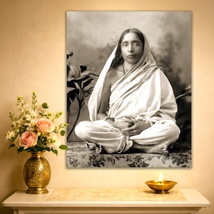 May include: Black and white portrait of a woman in traditional Indian clothing, seated with hands in her lap. The image is displayed on a wall, with a vase of flowers and a lit candle in the foreground.
