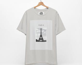 May include: A light gray t-shirt featuring a black and white image of the Eiffel Tower in Paris. The word "PARIS" is printed above the image. The shirt is hanging on a wooden hanger.