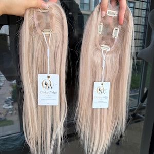 May include: Two light blonde hair toppers with straight, smooth strands. Each piece features a sheer base with metal pressure-sensitive clips attached for secure placement. A white tag with the text Olokey Wigs is attached to each hair topper.