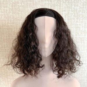 May include: A dark brown, curly wig displayed on a mannequin head. The wig has a black headband and a realistic hairline. The hair is styled in loose, voluminous curls, giving a full and textured look. The mannequin head is a neutral beige colour.