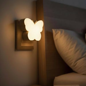 Op de afbeelding: Een vlindervormig nachtlampje is aangesloten op een stopcontact. Het licht geeft een zachte witte gloed, en de armatuur is gemonteerd op een houten basis. Op de achtergrond zijn een bed en een kussen te zien.