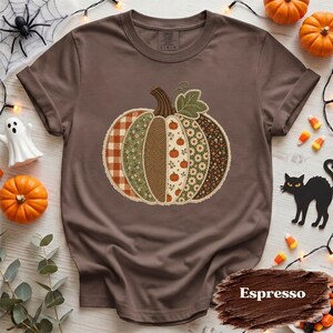 以下が含まれることがあります： 茶色の半袖Tシャツ。中央には、チェック柄、花柄、ヘリンボーン柄、小さなカボチャのプリントなど、さまざまなパターンの生地を組み合わせたカボチャの大きなグラフィックが描かれており、緑色の葉のアクセントが付いています。
