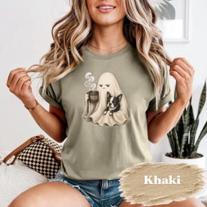 May include: A khaki t-shirt featuring a graphic of a ghost draped in a white sheet, holding a steaming mug labeled MEH, and sitting next to a black and white French Bulldog. The shirt has short sleeves and a crew neckline, displaying a casual fit.