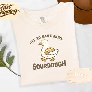 Può includere: T-shirt color avorio con un'anatra dei cartoni animati che trasporta una pagnotta. Il testo sulla maglietta recita "OFF TO BAKE MORE SOURDOUGH". La maglietta è del marchio Comfort Colors. Una grafica "Fast Shipping" è nell'angolo in alto a sinistra.