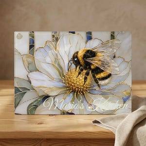 May include: A rectangular glass cutting board featuring a stained-glass style illustration of a fuzzy bumblebee resting on a white flower with gold outlines. The text Maria's Kitchen is printed in white cursive script across the bottom center.