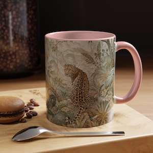 May include: A ceramic mug with a soft pink interior and handle, featuring a detailed wrap-around illustration of a leopard sitting in a lush, muted-tone jungle environment with foliage and trees, set against a neutral, textured background.