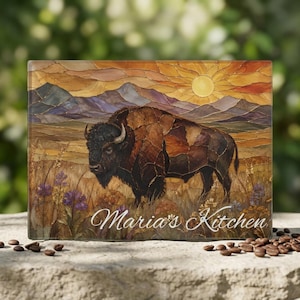 May include: A rectangular glass cutting board featuring a stained-glass style illustration of a brown bison standing in a field of tall grass and purple wildflowers, with mountains and a sun in the background. The text Maria's Kitchen is printed.
