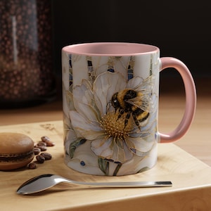 May include: A ceramic mug with a pastel pink interior and handle, featuring a stained-glass style illustration of a bumblebee resting on a large white flower with gold outlines. The design includes subtle blue and green accents within the petals.