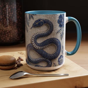 May include: A ceramic mug featuring a detailed blue snake illustration coiled against a textured light grey background with blue floral accents. The mug has a solid light blue interior and a matching light blue handle, providing a vibrant contrast.