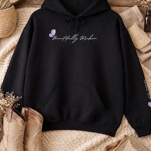 Op de afbeelding: Zwarte hoodie met een voorzak, met de tekst "Beautifully Broken" en een vlindergrafiek. De mouw heeft extra tekst en een vlinder. De afbeelding bevat ook een paar beige cowboylaarzen.