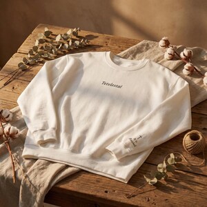 May include: Cream-colored sweatshirt with the word "Tetelestai" printed across the chest. The right sleeve has the text "It is Finished" with a cross symbol. The sweatshirt is displayed on a wooden surface with decorative elements.