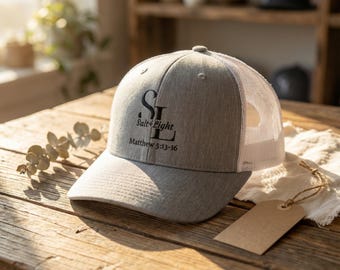 May include: A gray trucker hat with a white mesh back. The hat features the text "Salt + Light" and "Matthew 5:13-16" in black. The hat is sitting on a wooden surface with a blank tag.