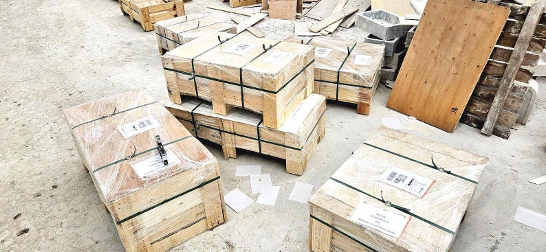 May include: Several wooden crates, wrapped in clear plastic and secured with black straps, are stacked on a concrete surface. Some crates are stacked on top of each other. Various pieces of wood and other construction materials are scattered around the crates.