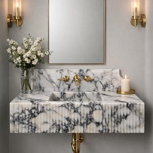 May include: A marble bathroom vanity unit with a built-in sink and gold-coloured taps and waste pipe. The countertop has a white and grey veined pattern. A mirror hangs above the sink, and wall sconces flank it. A vase of flowers sits to the left.