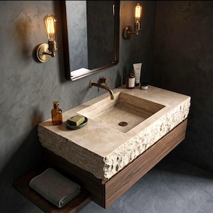 May include: A bathroom vanity featuring a rectangular beige travertine stone sink with a rough, chiseled front edge. The sink is mounted on a dark wood cabinet with a matching shelf below. It includes a brass faucet, two wall-mounted Edison bulb lights, a mirror, and various toiletries.