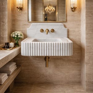 May include: A stylish bathroom interior with a white marble sink with fluted detailing and gold fixtures. An arched mirror hangs above the sink, flanked by two sconces. The room is decorated in neutral tones, with beige walls and flooring.