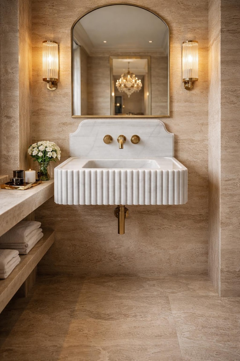 May include: A luxurious bathroom scene featuring a white marble sink with fluted detailing, gold fixtures, and a matching arched mirror. Two elegant sconces flank the mirror. The room has a neutral color palette with beige walls and flooring.