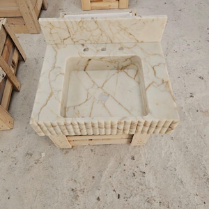 Pu&ograve; includere: Un mobile da bagno rettangolare in marmo con lavabo integrato. La pietra bianco sporco presenta venature marroni. Il bordo anteriore del mobile ha un design a coste. Sono presenti due fori per i rubinetti. Il mobile &egrave; appoggiato su un bancale di legno.