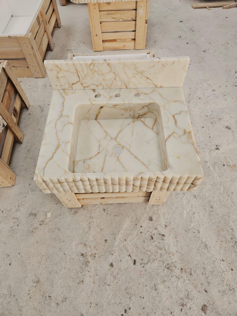Pu&ograve; includere: Un mobile da bagno rettangolare in marmo con lavabo integrato. La pietra bianco sporco presenta venature marroni. Il bordo anteriore del mobile ha un design a coste. Sono presenti due fori per i rubinetti. Il mobile &egrave; appoggiato su un bancale di legno.