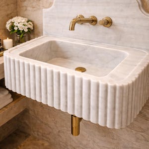 May include: A white marble bathroom sink with a fluted design. The sink has a rectangular basin and is paired with gold-colored fixtures, including a faucet and drain. The sink is mounted on a wall with a matching marble backsplash.