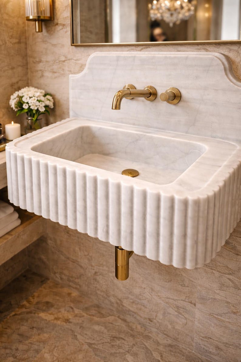 May include: A white marble bathroom sink with a fluted design. The sink has a rectangular basin and is paired with gold-colored fixtures, including a faucet and drain. The sink is mounted on a wall with a matching marble backsplash.