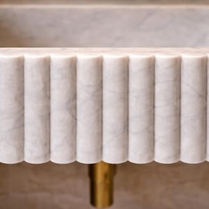 May include: Close-up of a white marble sink with a fluted, vertical design. The sink has a rectangular basin and a decorative, scalloped edge. A gold-colored drain pipe is visible below the sink.