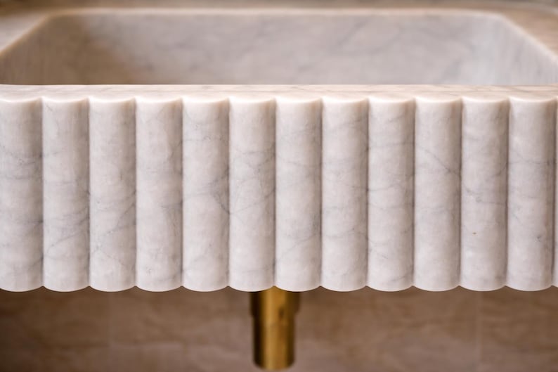 May include: Close-up of a white marble sink with a fluted, vertical design. The sink has a rectangular basin and a decorative, scalloped edge. A gold-colored drain pipe is visible below the sink.