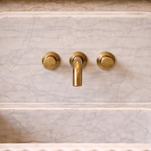 May include: A close-up of a marble sink with a brass faucet and knobs. The sink is rectangular with a curved front edge. The faucet is centered above the sink, with two knobs on either side. The marble has gray veining.