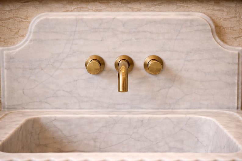 May include: A close-up of a marble sink with a brass faucet and knobs. The sink is rectangular with a curved front edge. The faucet is centered above the sink, with two knobs on either side. The marble has gray veining.