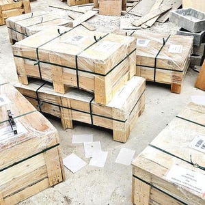 K&ouml;nnte beinhalten: Mehrere Holzkisten, umwickelt mit durchsichtigem Kunststoff und mit schwarzen Gurten gesichert, sind auf einer Betonoberfl&auml;che gestapelt. Einige Kisten sind &uuml;bereinander gestapelt. Verschiedene Holzst&uuml;cke und andere Baumaterialien liegen herum.