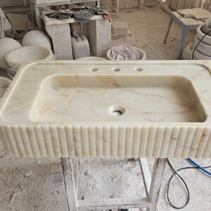 K&ouml;nnte beinhalten: Ein rechteckiges Waschbecken aus hellbeige Marmor mit feinen Maserungen. Es hat ein zentrales Abflussloch und drei L&ouml;cher f&uuml;r die Wasserh&auml;hne. Die Vorderseite weist vertikale Rippendetails auf. Weitere Steinbecken und Kugeln sind im Hintergrund zu sehen.