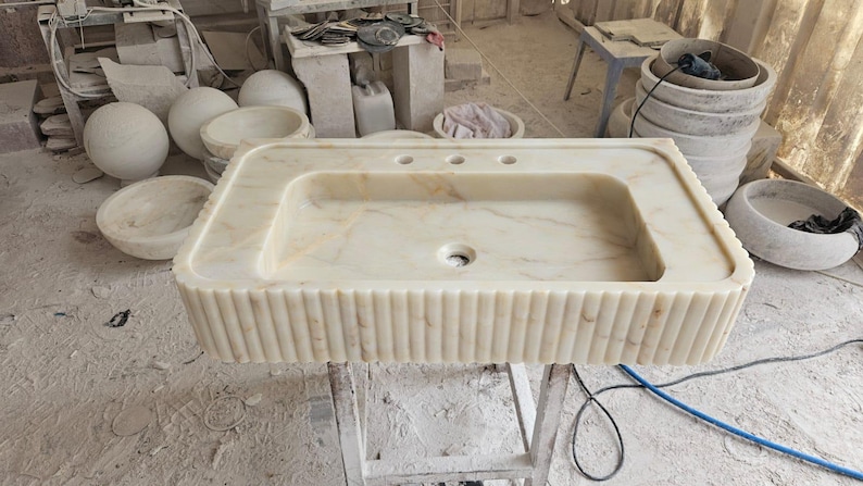 K&ouml;nnte beinhalten: Ein rechteckiges Waschbecken aus hellbeige Marmor mit feinen Maserungen. Es hat ein zentrales Abflussloch und drei L&ouml;cher f&uuml;r die Wasserh&auml;hne. Die Vorderseite weist vertikale Rippendetails auf. Weitere Steinbecken und Kugeln sind im Hintergrund zu sehen.