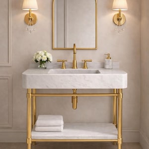 Puede incluir: Un lujoso mueble de baño con lavabo de mármol y accesorios dorados. El mueble tiene un marco dorado, una balda para toallas y un espejo arqueado a juego. Dos apliques con pantallas plisadas flanquean el espejo. Un jarrón con flores blancas.