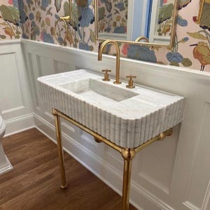 Pode incluir: Um lavat&oacute;rio retangular de m&aacute;rmore branco com um design exterior canelado, suportado por uma estrutura de metal dourado polido. O lavat&oacute;rio possui uma torneira dourada a condizer com dois man&iacute;pulos separados, montada na superf&iacute;cie.