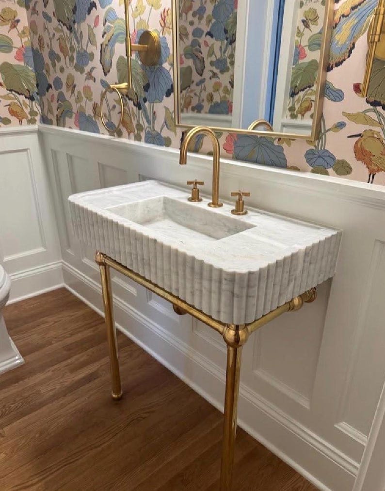 Pode incluir: Um lavat&oacute;rio retangular de m&aacute;rmore branco com um design exterior canelado, suportado por uma estrutura de metal dourado polido. O lavat&oacute;rio possui uma torneira dourada a condizer com dois man&iacute;pulos separados, montada na superf&iacute;cie.