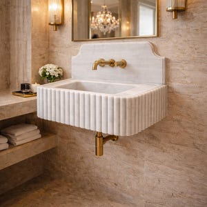 May include: A white marble sink with a fluted design and gold fixtures. The sink is mounted on a wall with a matching marble backsplash. Two gold sconces flank a large arched mirror. The bathroom has a luxurious, spa-like aesthetic.