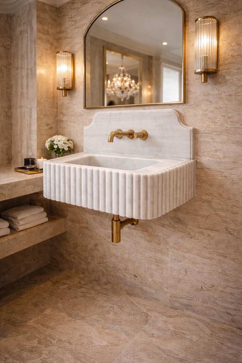 May include: A white marble sink with a fluted design and gold fixtures. The sink is mounted on a wall with a matching marble backsplash. Two gold sconces flank a large arched mirror. The bathroom has a luxurious, spa-like aesthetic.
