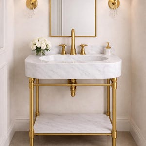 May include: A luxurious bathroom vanity with a marble sink and gold-toned fixtures. The sink is supported by a gold metal frame with a lower shelf. A gold-framed mirror and sconces with pleated shades complete the elegant design.
