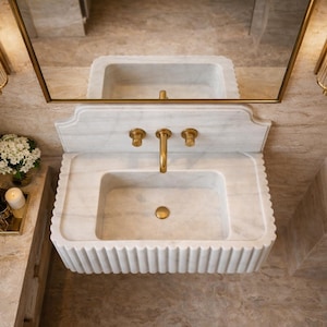 May include: A luxurious marble sink with gold fixtures, including a faucet and two handles. The sink has a rectangular basin and a fluted apron. Two gold sconces flank a gold-framed mirror above the sink. A small tray with a candle and flowers sits to the side.
