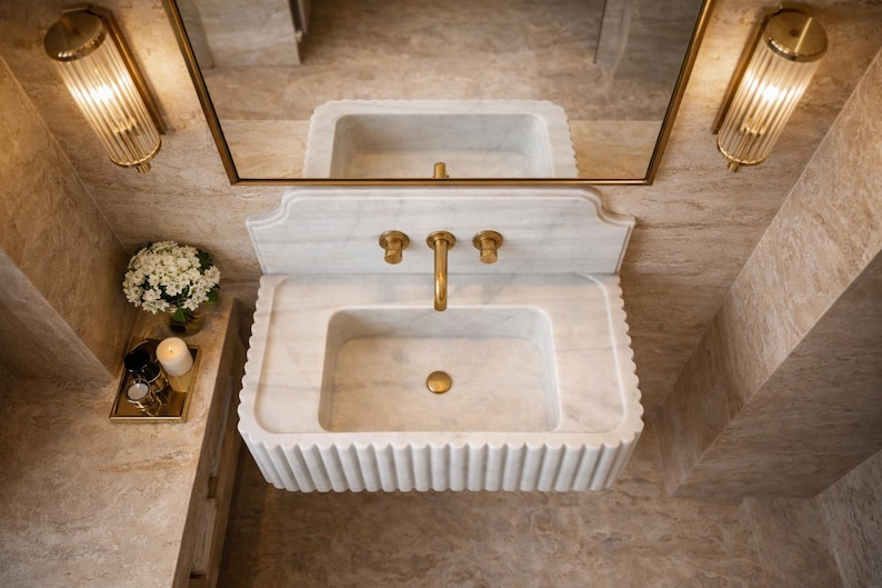 May include: A luxurious marble sink with gold fixtures, including a faucet and two handles. The sink has a rectangular basin and a fluted apron. Two gold sconces flank a gold-framed mirror above the sink. A small tray with a candle and flowers sits to the side.